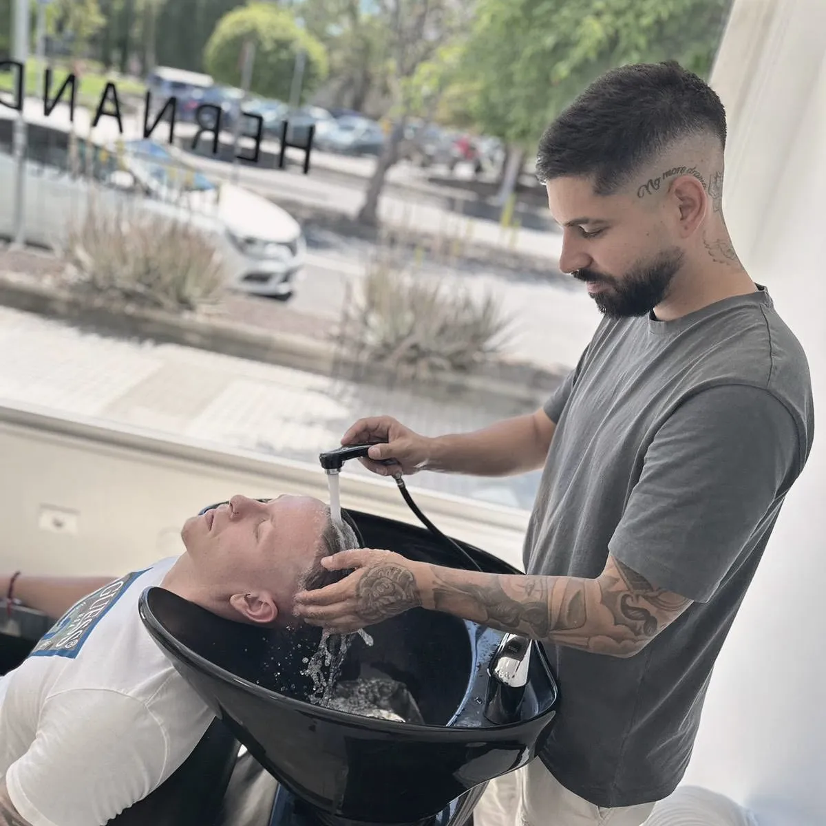 Hombre recibiendo un lavado de cabello en un salón de belleza, mientras un estilista aplica agua sobre su cabeza. Se observa un ambiente moderno y luminoso.