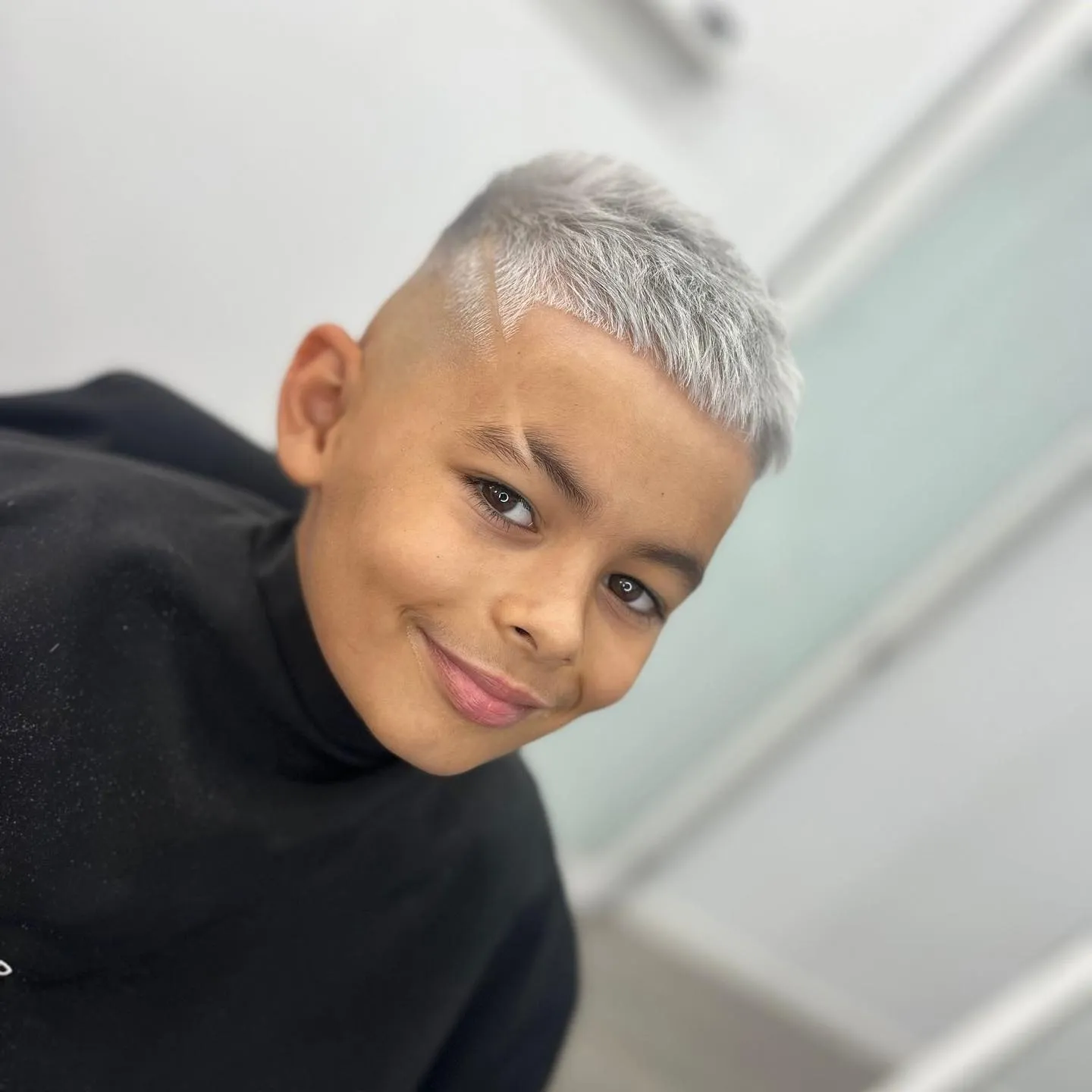 Niño con cabello corto y rubio platinado, sonriendo mientras está en una barbería. Lleva una capa negra y tiene un diseño en el costado de la cabeza. Fondo desenfocado de un salón moderno.